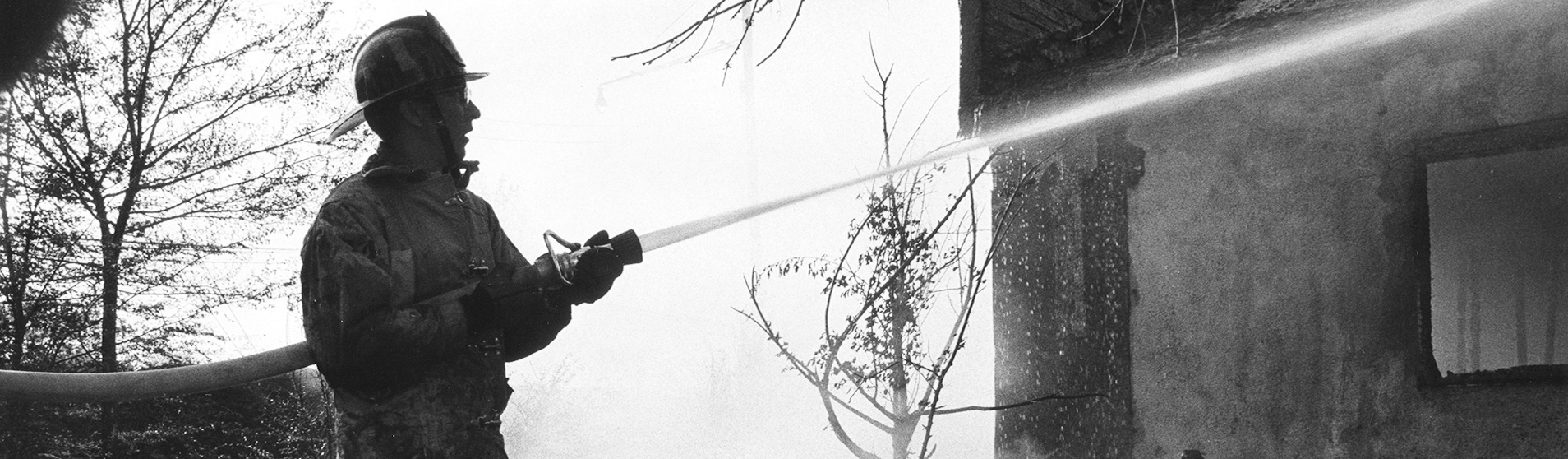 Black and white photo of a Greeley firefighter battling a burning building with a fire hose