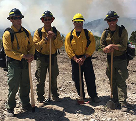 Four firefighters standing outside near a smoldering wildfire