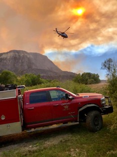 Fire rescue truck parked on a grassy mountainside with a helicopter flying overhead against a cloudy sky